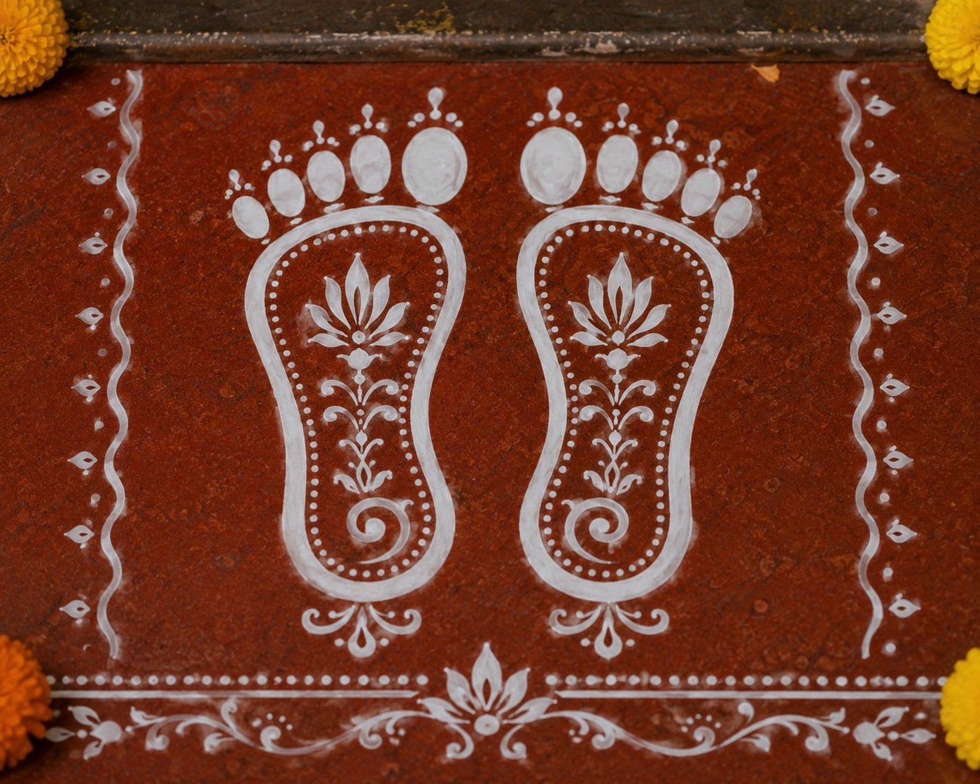 Close-up of two Lakshmi feet in white rice paste, each sole filled with a lotus and dotted outline, on red geru