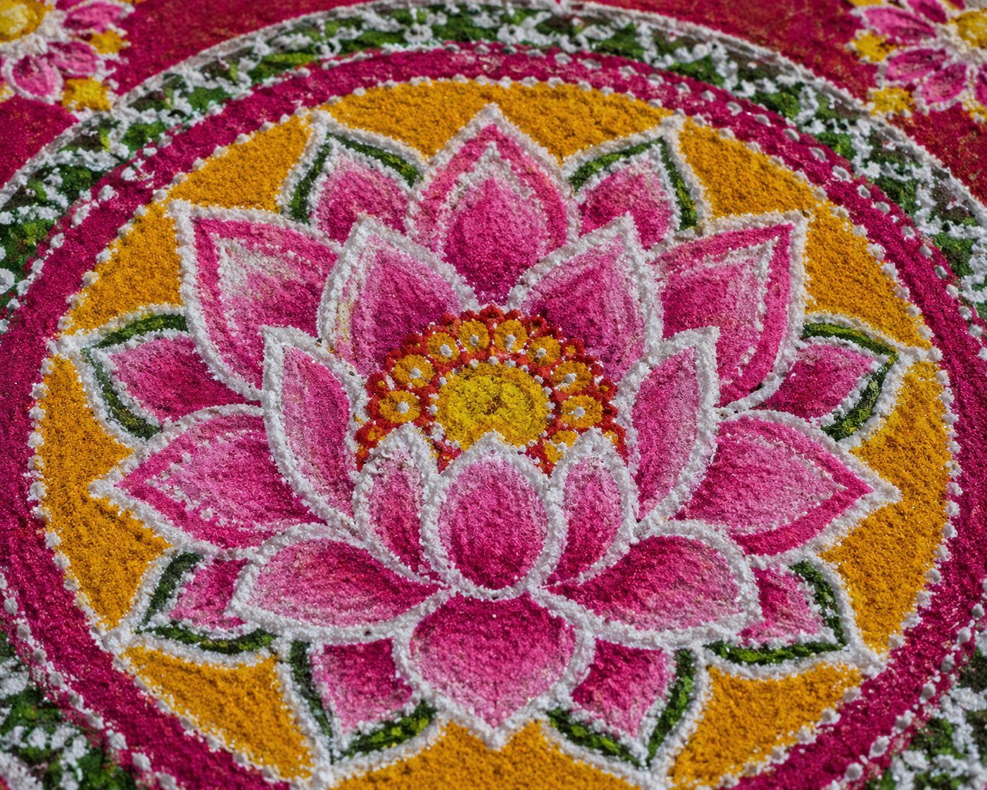 Close-up of a pink and orange lotus drawn in coloured powder rangoli, petals outlined in white, at the centre of a Saraswati chowki